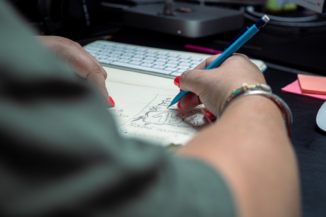 Une main esquisse un logo au crayon sur un carnet, a cote d'un clavier.