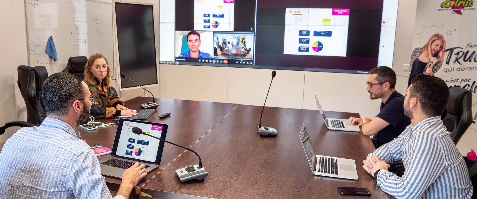 L'équipe au travail autour de la grande table, face à l'écran de travail et à un intervenant en visio.