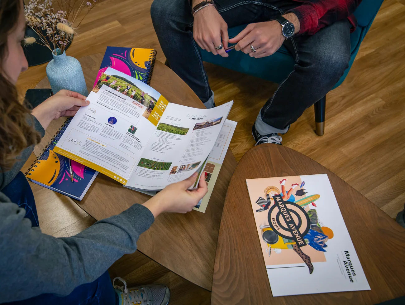 Une personne feuillette une brochure imprimée sur une table basse, à côté d'autres documents et d'une affiche.