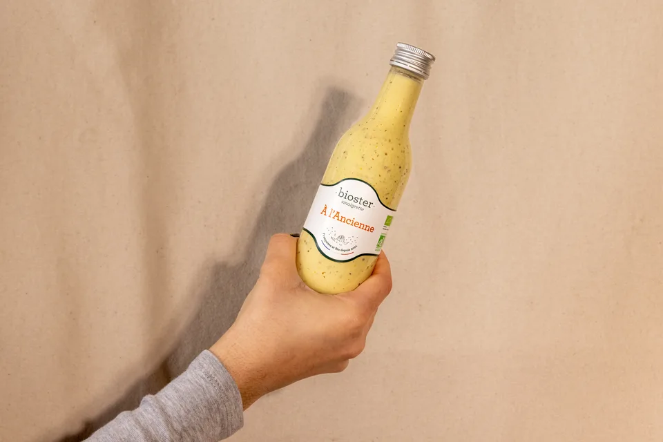 Une bouteille de vinaigrette Bioster tenue en main sur fond beige.