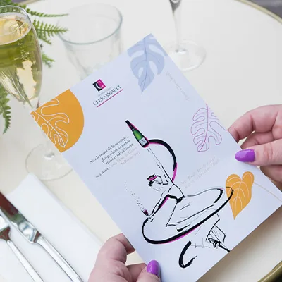 Une carte Champagne Clérambault illustrée, tenue à la main près de deux flûtes servies.