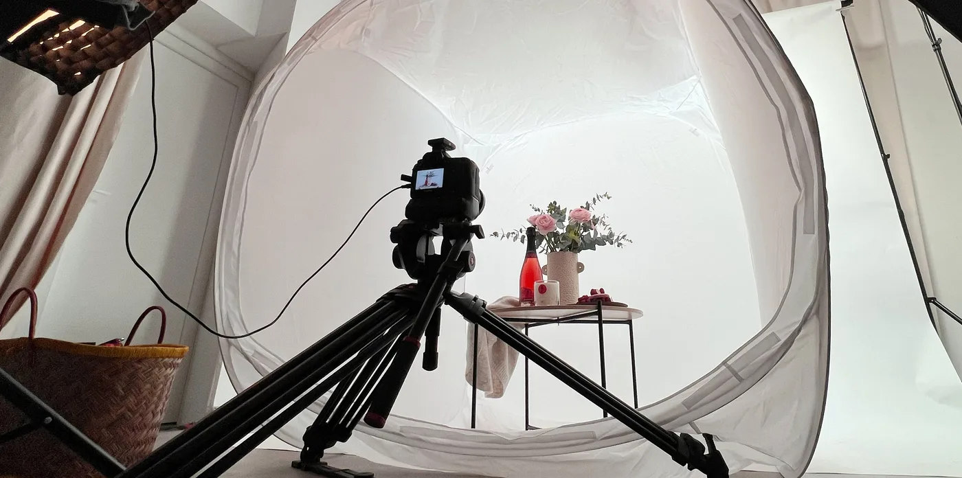 Une séance de production en studio : une caméra sur trépied filme une bouteille et un bouquet dans une tente de lumière.